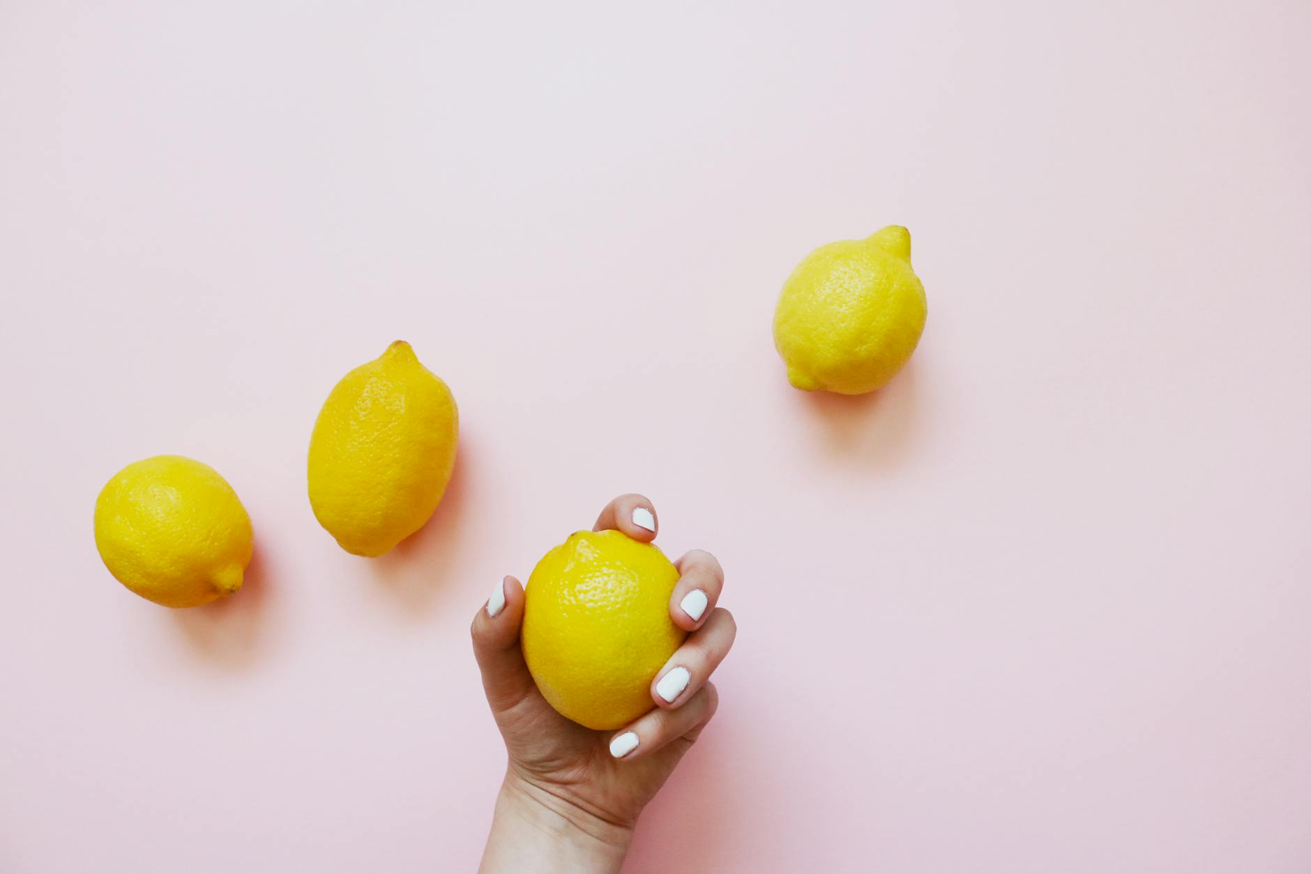 Eine Hand mit weißem Nagellack hält eine Zitrone vor einem weichen rosa Hintergrund, umgeben von drei weiteren Zitronen.