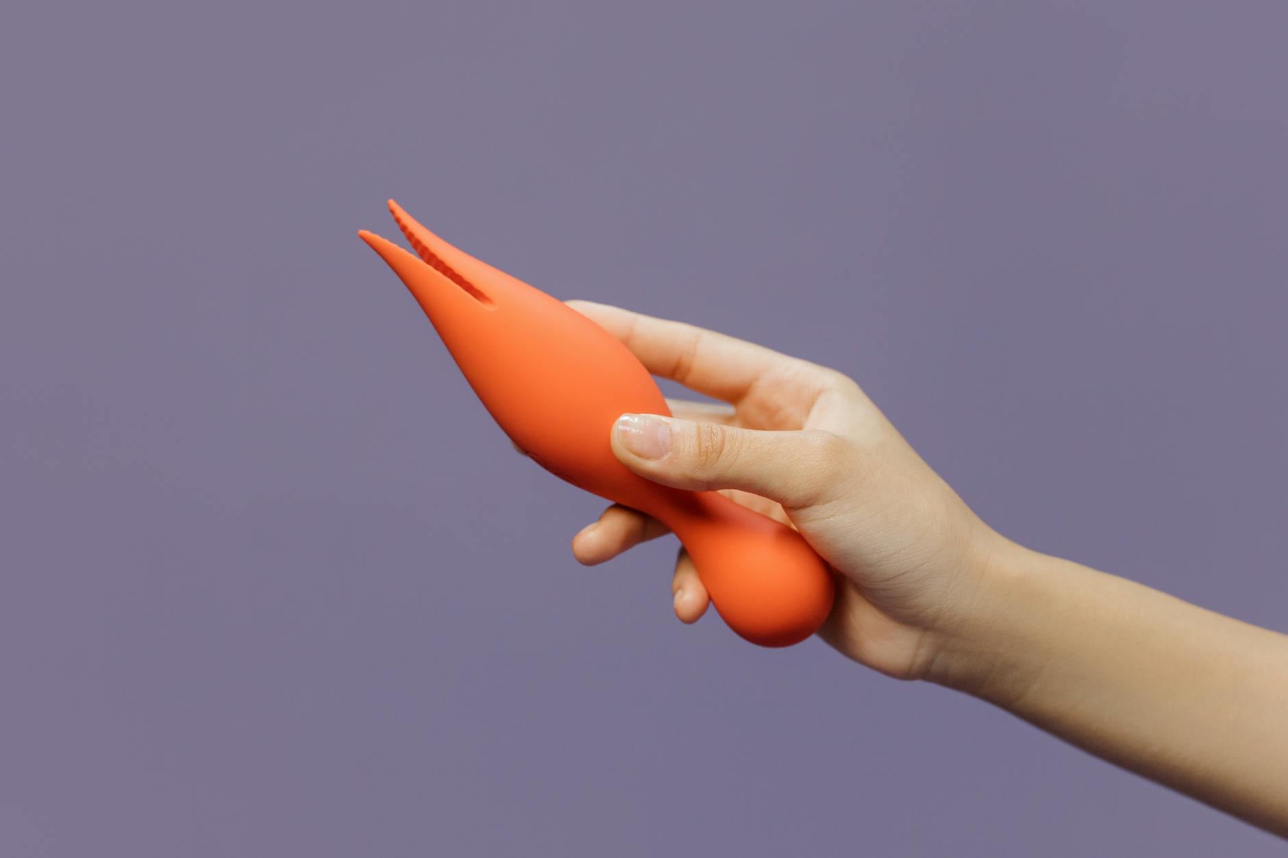 Eine Hand hält einen orangefarbenen Vibrator gegen minimalistischen violetten Hintergrund, zeigt moderne Sinnlichkeit